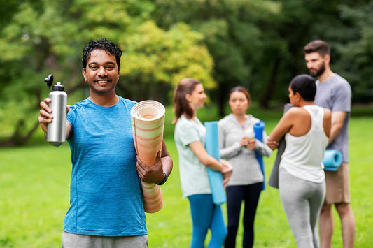 Smiling man holding a reusable water bottle and a rolled yoga mat stands in a park, with a small group of people chatting in the background, some holding yoga mats.